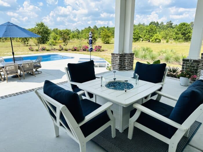 4 white chairs with blue cushions surrounding a white fire table on the back porch with the pool in the background